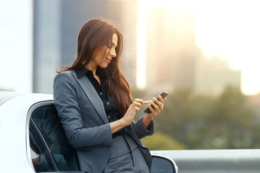 Een vrouw in zakelijke kleding leunt tegen een auto en gebruikt haar smartphone. Carano biedt met LAPID Fleet een mobiele oplossing voor de digitale controle van rijbewijzen. De app maakt flexibele controles onderweg mogelijk, vermindert de administratieve rompslomp en voldoet op betrouwbare wijze aan de wettelijke vereisten voor het wagenpark.  